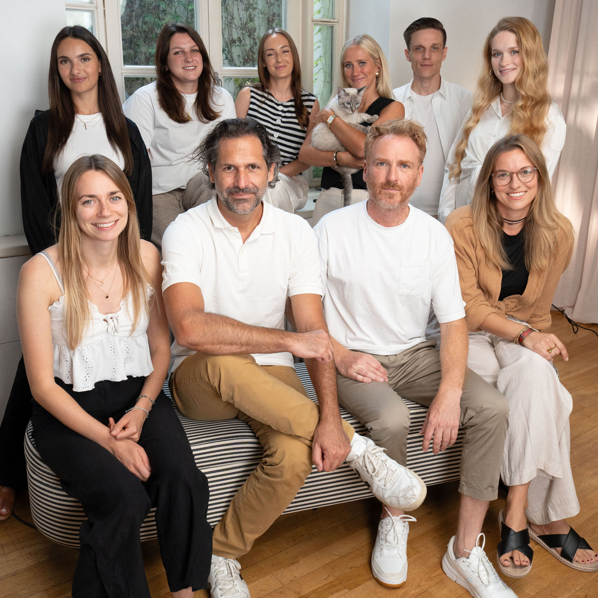 Das LucyBalu-Team 2025: zehn lächelnde Personen in heller Kleidung in einem Raum mit großen Fenstern, zwei davon halten Siamkatzen auf dem Arm.
