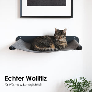 Getigerte Katze liegt zusammengerollt auf einer grauen Hängematte aus Wollfilz, die unter einem gerahmten Bild an der Wand montiert ist.
