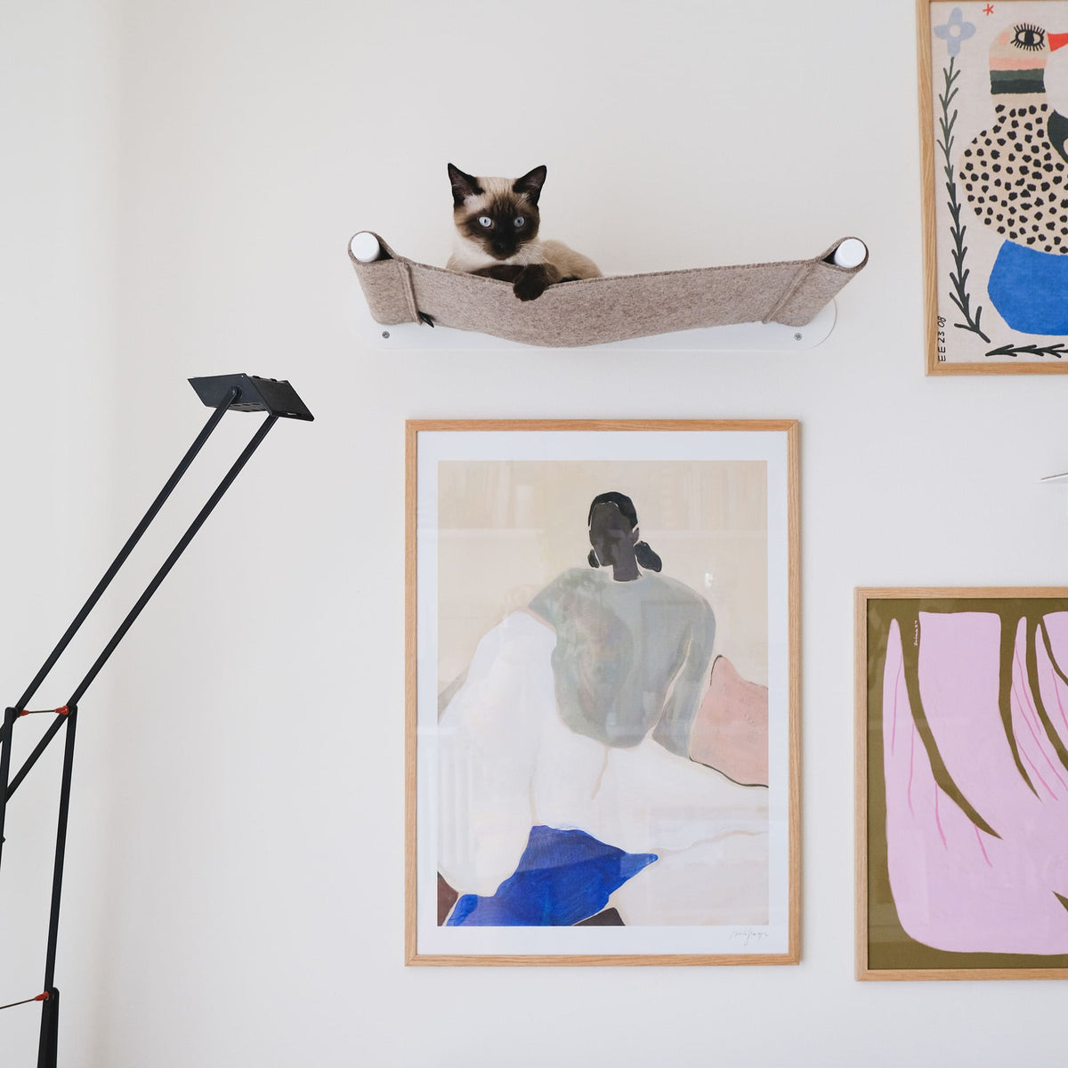 Siamkatze sitzt in einer LucyBalu Wand-Hängematte aus hellbraunem Filz mit weißer Halterung, darunter gerahmte Kunstdrucke an einer hellen Wand und eine schwarze Stehlampe.